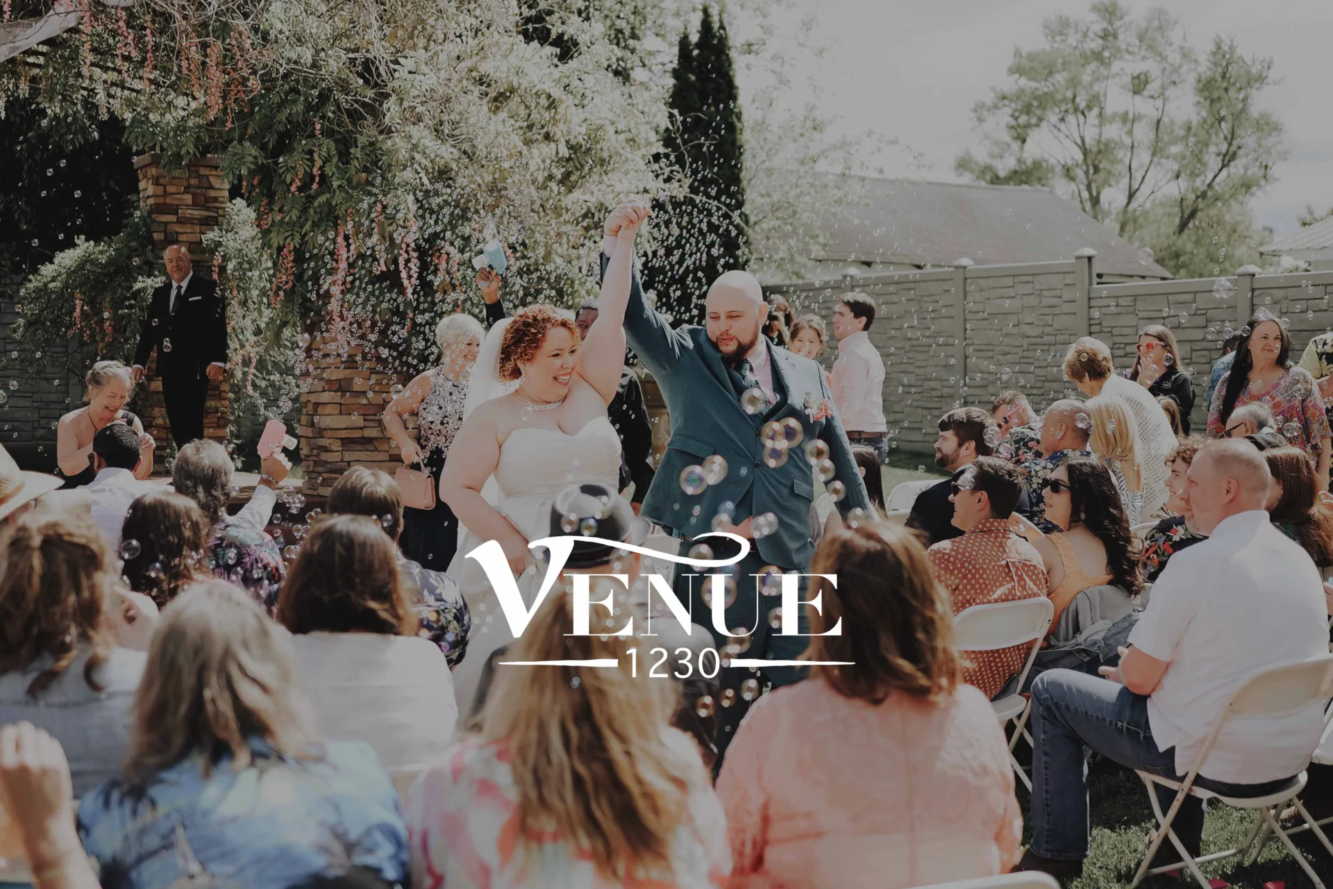 Outdoor wedding reception: bride and groom dancing amid floating bubbles while guests cheer and watch in a sunny backyard setting.