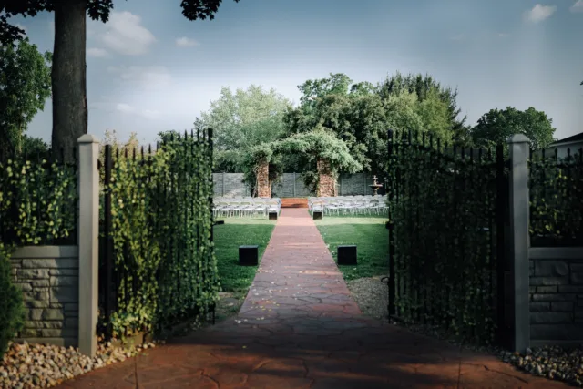 Brick pathway leads to an outdoor ceremony area with white folding chairs on both sides and lush trees in the background.