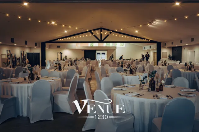 Banquet hall prepared for a formal event with round tables, white chair covers, and warm string lights overhead; a logo reads 'Venue 1230' at the front edge of the scene.