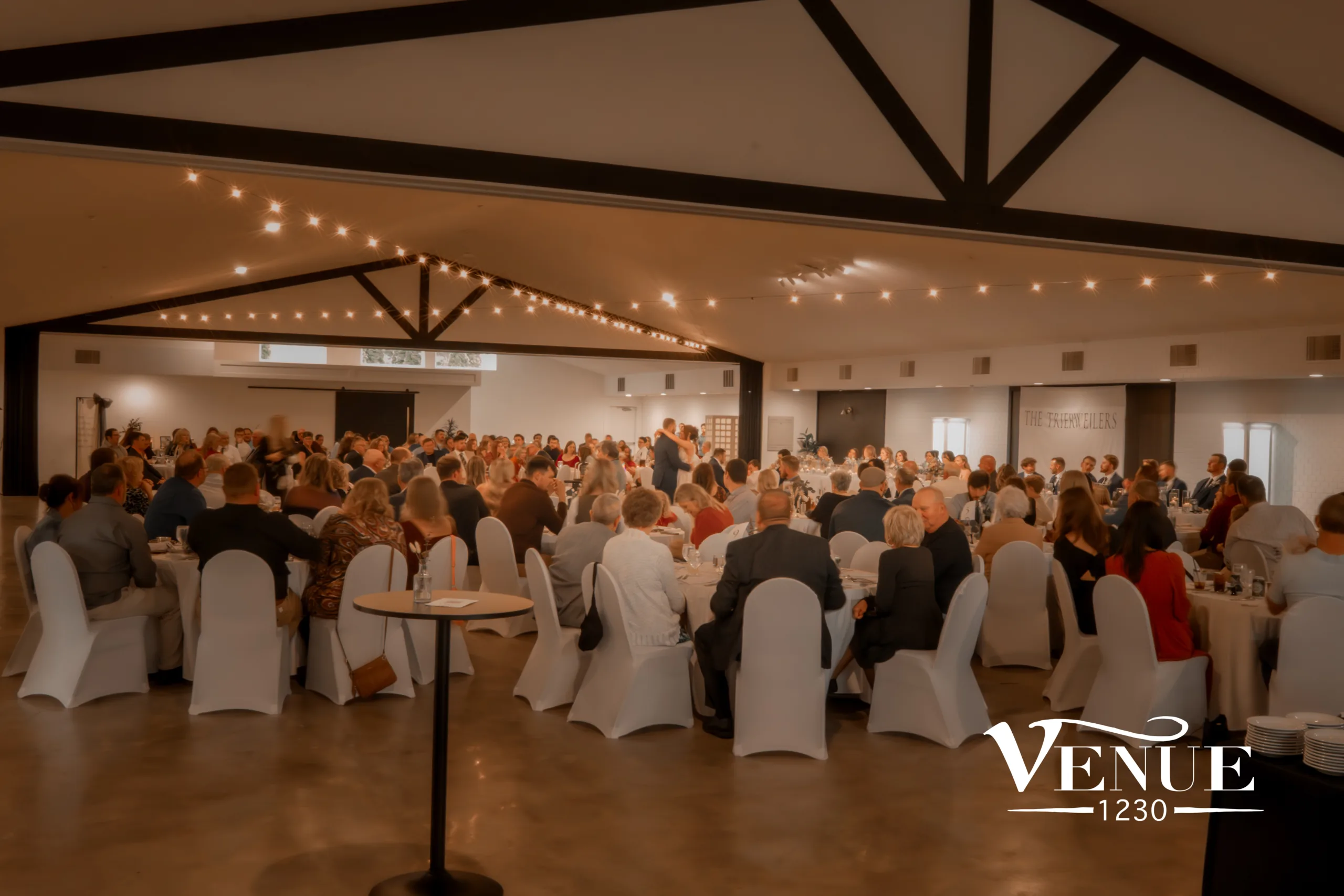 Banquet hall filled with round tables and seated guests at a formal reception under warm string lights.