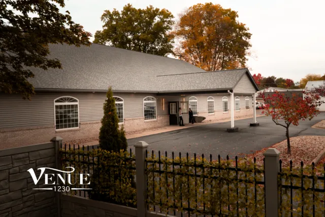 Exterior view of a venue building with arched windows, a slate roof, and autumn trees—people stand near the entrance; text 'Venue 1230' visible at the bottom left.